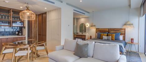 A modern hotel room with a sofa, dining table, chairs, a bed with pillows, a bedside table, a lamp, and a large window.