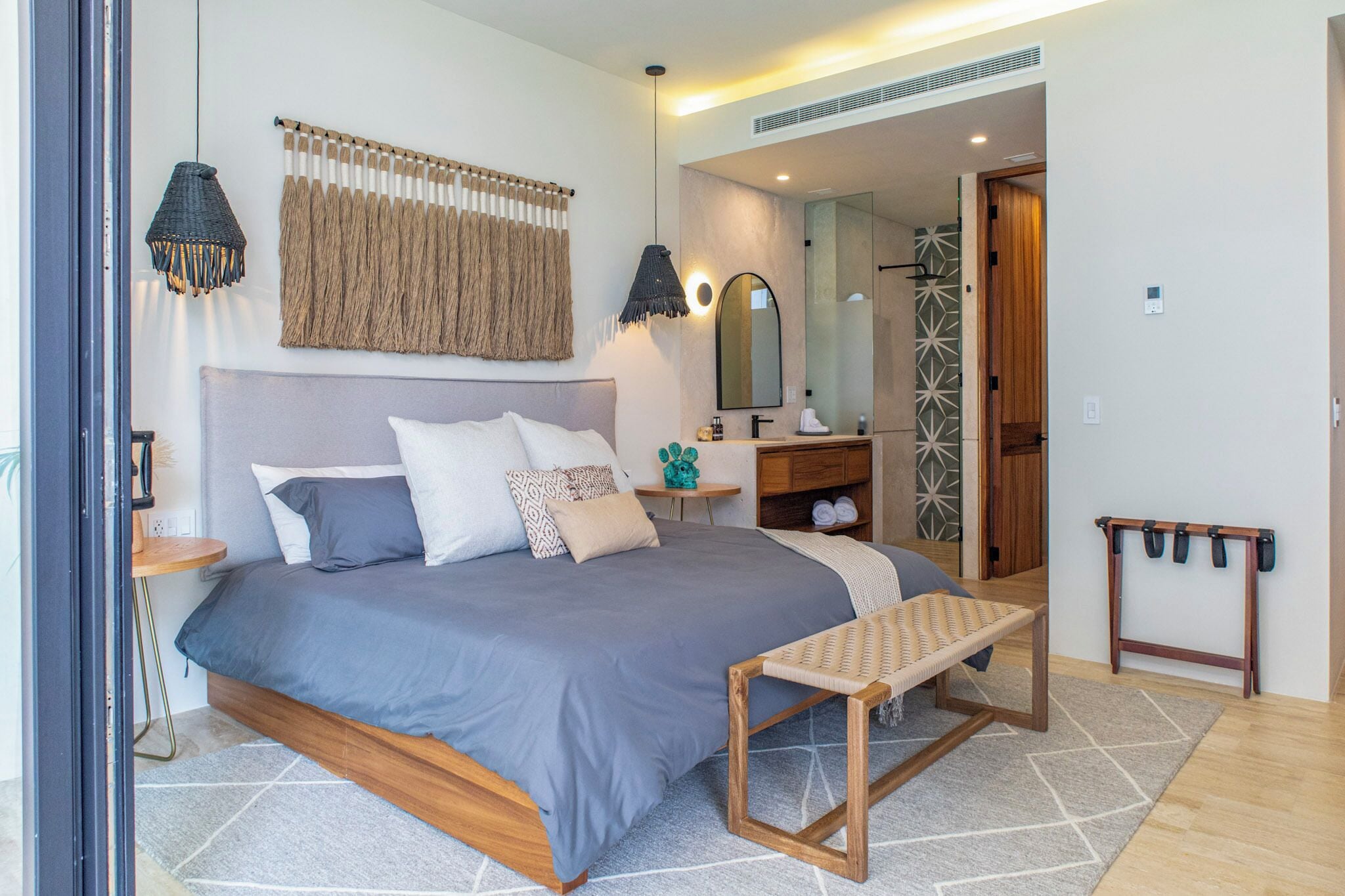 A modern bedroom with a large bed, bedside tables, a bench, a mirror, and a bathroom visible through an open door.