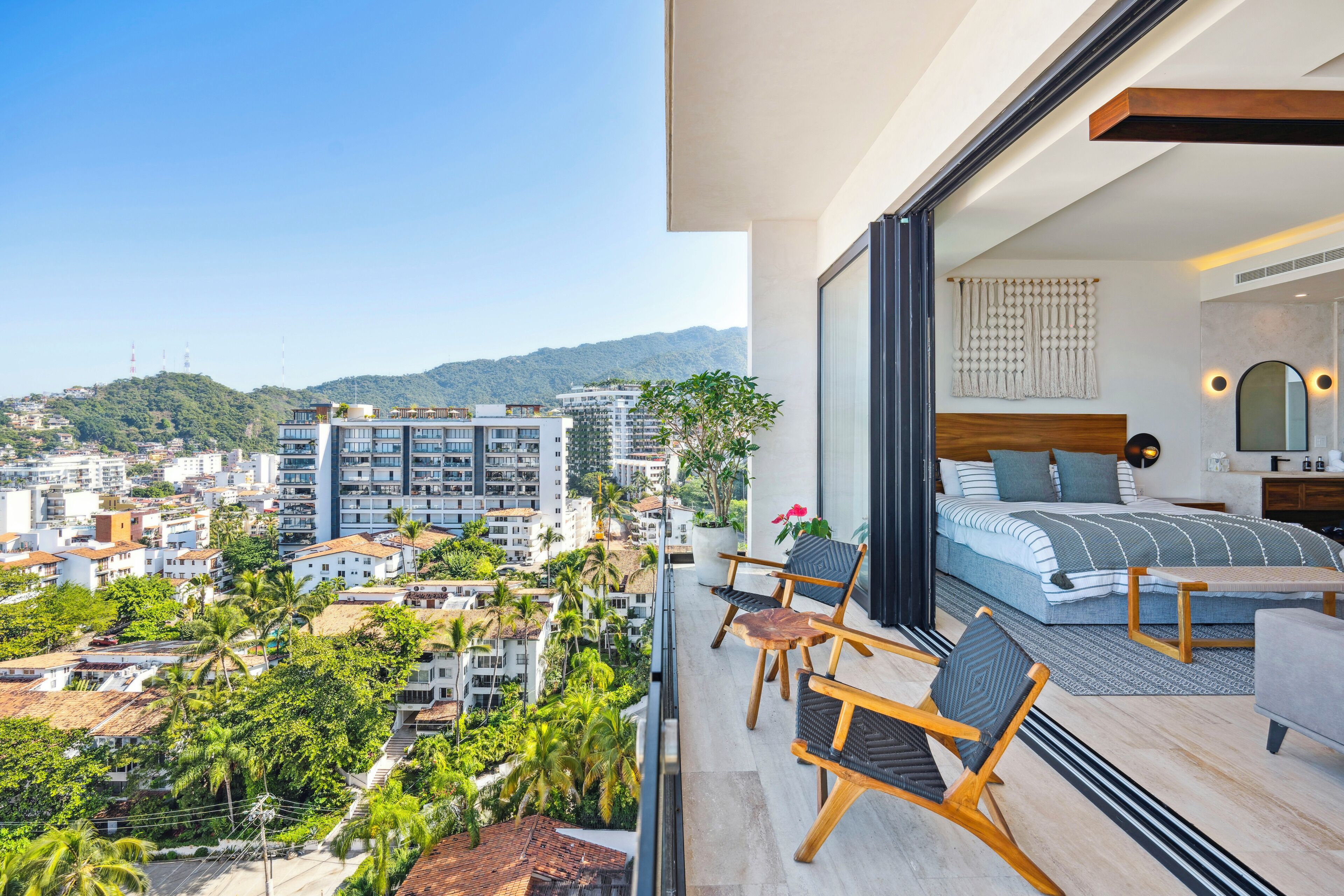 A balcony with a bed, two chairs, and a view of a cityscape and mountains.