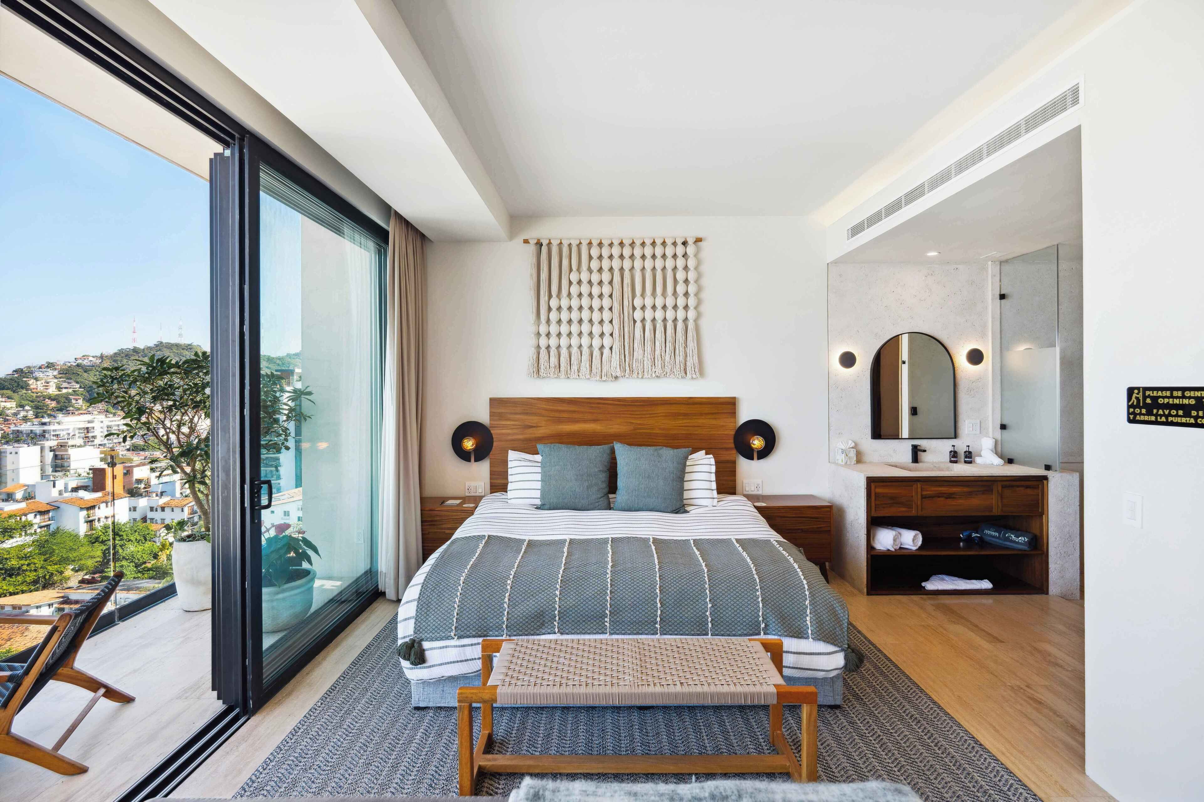 Una habitación de hotel moderna con una cama grande, una mesita de noche, un banco, un baño con espejo y vistas a la ciudad.