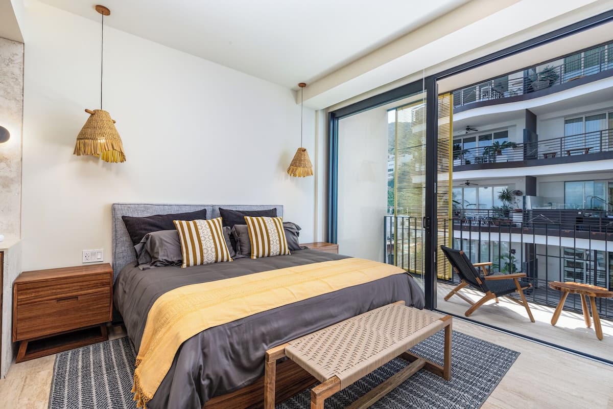 A modern bedroom with a large bed, bedside tables, a bench, and a balcony with outdoor seating.