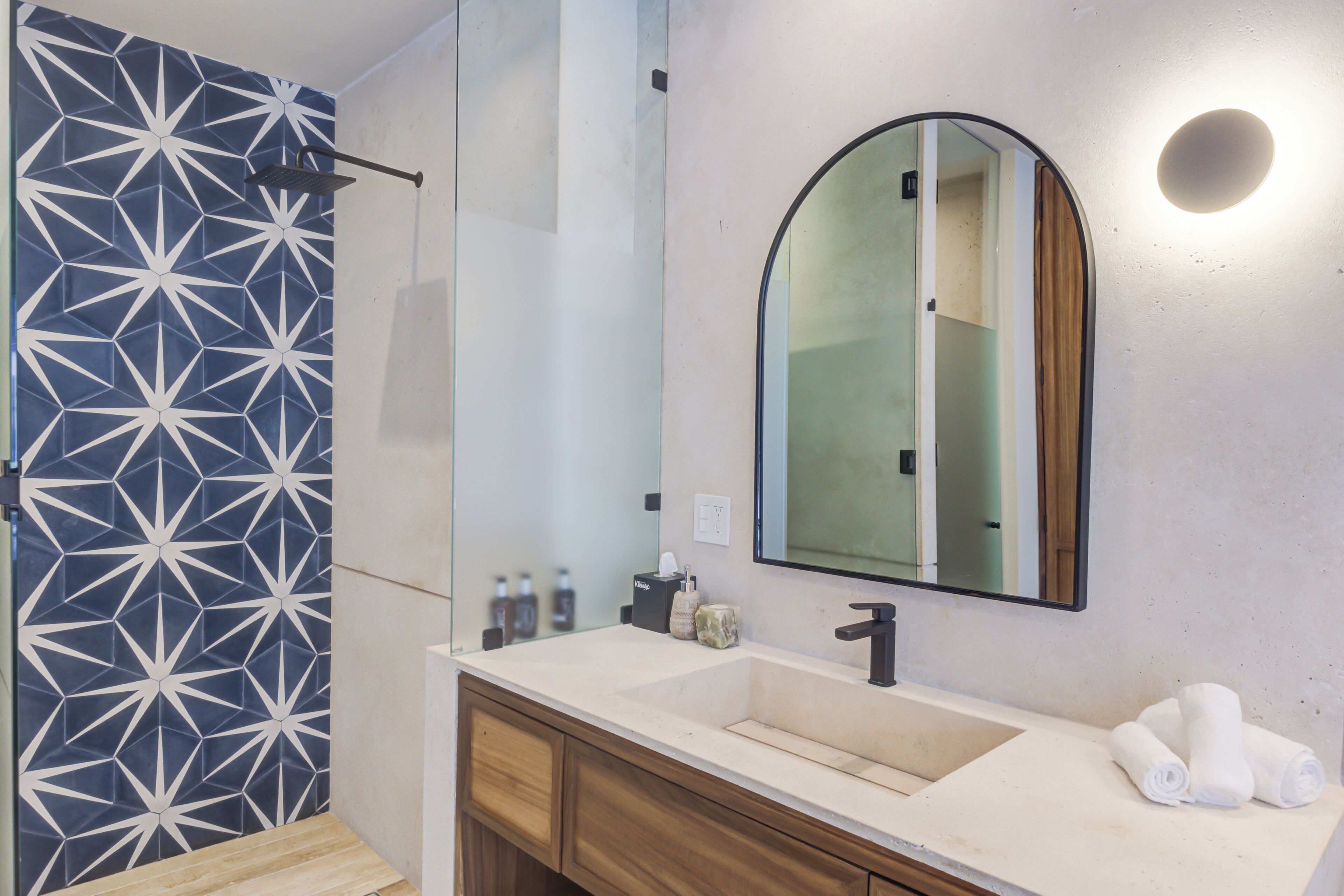 Une salle de bain moderne avec un mur recouvert de carreaux aux motifs géométriques, un grand miroir et un lavabo blanc agrémenté de meubles en bois.