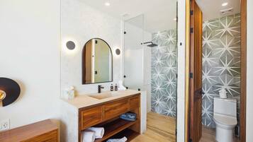 Un baño moderno con tocador de madera, un espejo grande, ducha con azulejos de diseño geométrico y un inodoro.