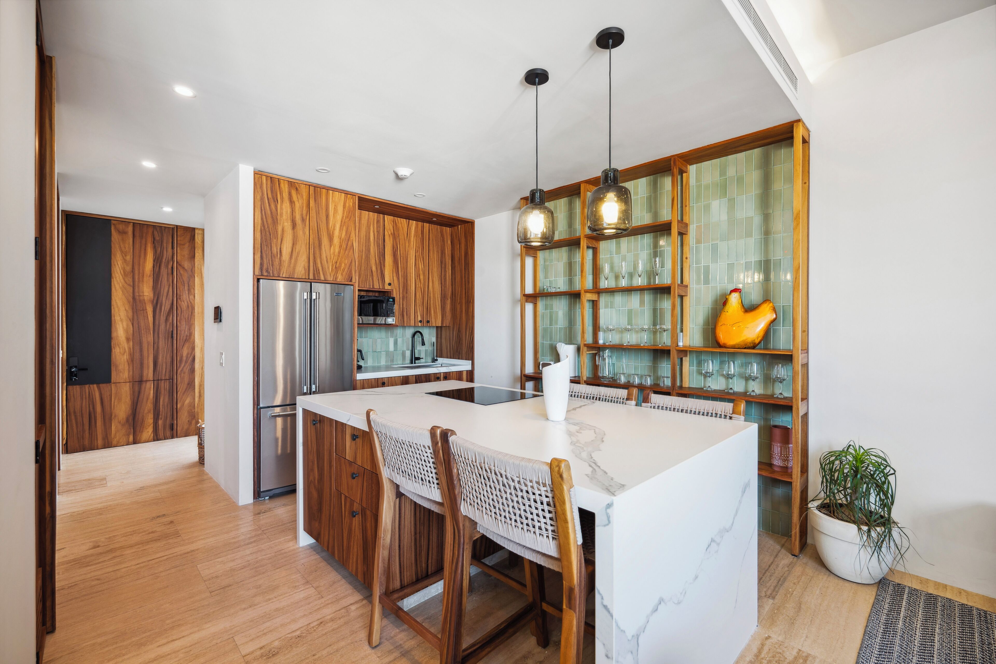 A modern kitchen with wooden cabinets, a stainless steel refrigerator, a marble island, and pendant lighting.