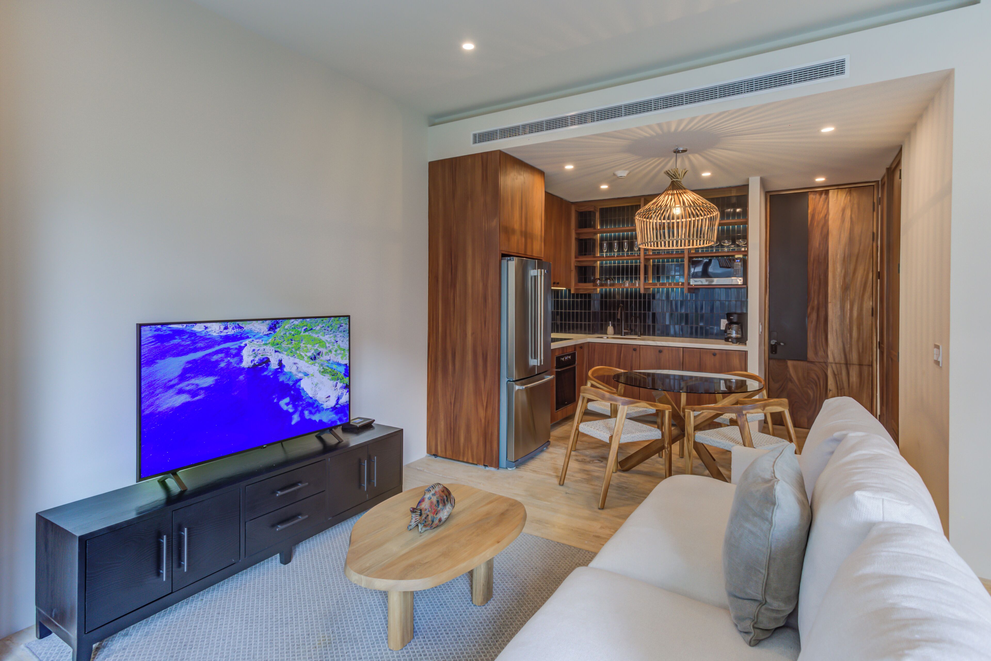 A modern living room with a flat-screen TV, wooden furniture, and a kitchen area with wooden cabinets and a built-in oven.