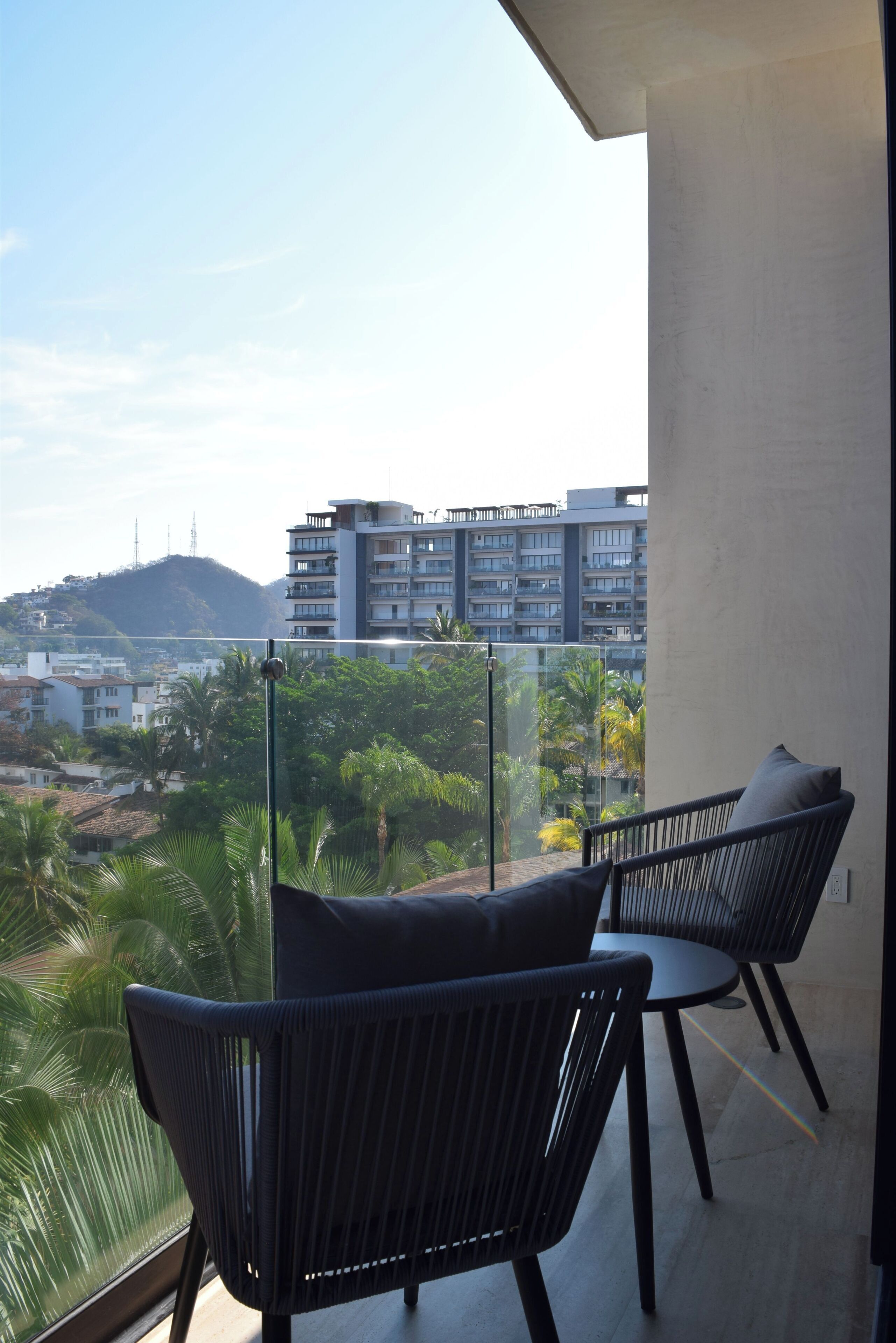 Un balcón con dos sillas y una barandilla de vidrio, con vistas a un paisaje urbano de edificios y palmeras.
