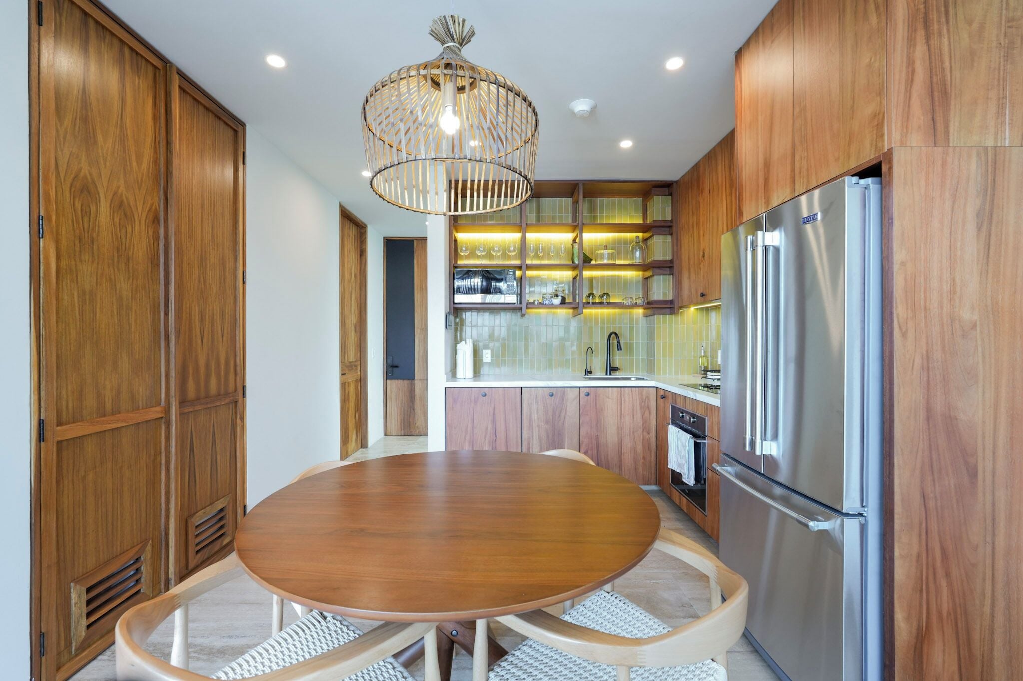 A modern kitchen with wooden cabinets, a stainless steel refrigerator, a round dining table, and a hanging light fixture.