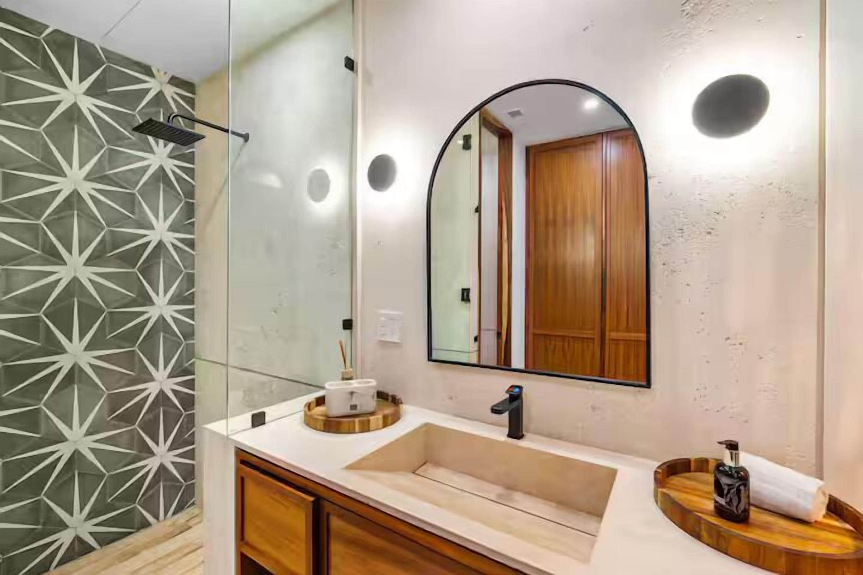 A modern bathroom with a geometric-patterned wall, a large mirror, a sink, and wooden cabinetry.