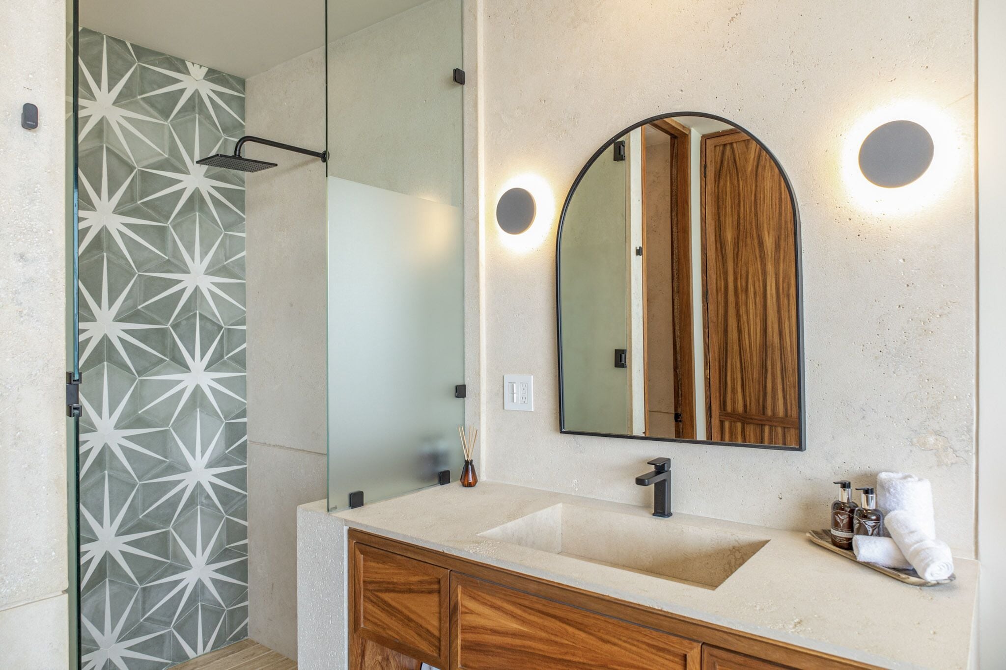 A modern bathroom with a geometric-patterned shower wall, a large mirror with a wooden frame, and a vanity with a beige countertop.