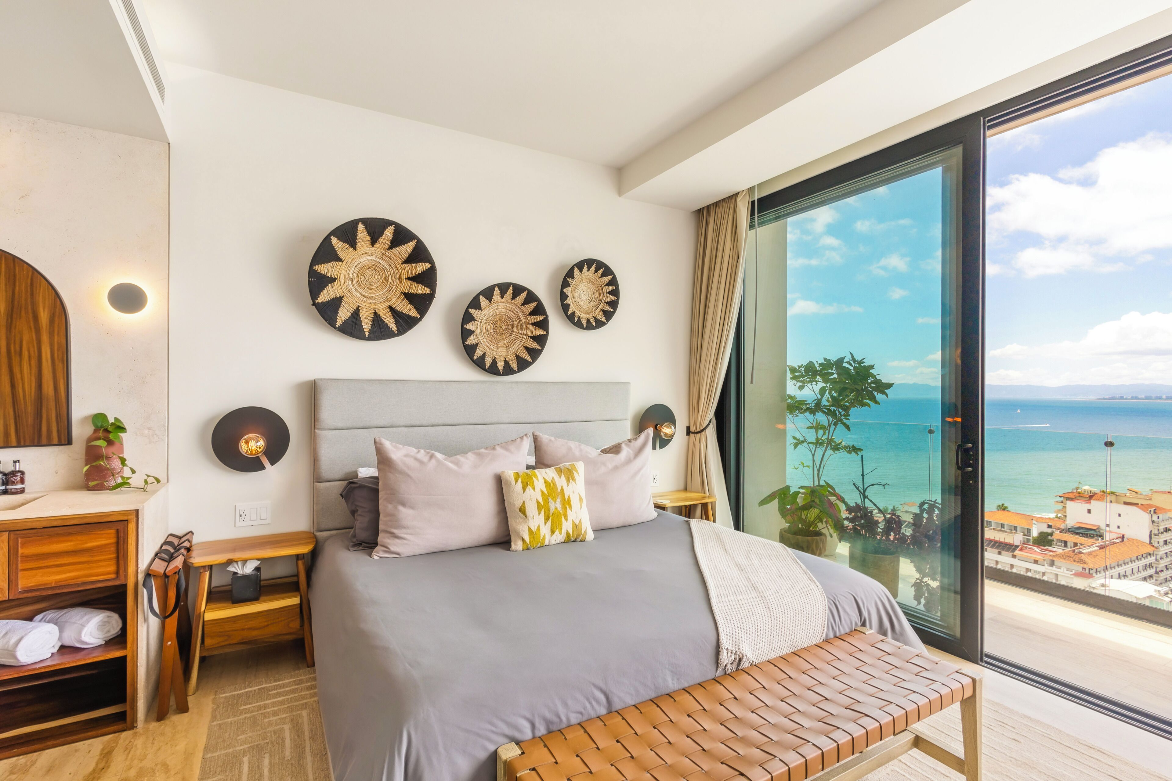 A modern bedroom with a large bed, bedside tables, a wooden bench, and a view of the sea.