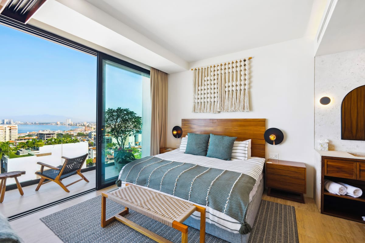 A modern bedroom with a large bed, a view of the city, and a balcony with a chair.