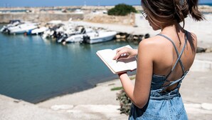 Une personne est assise au bord de l’eau, en train de lire un livre, avec des bateaux amarrés en arrière-plan.