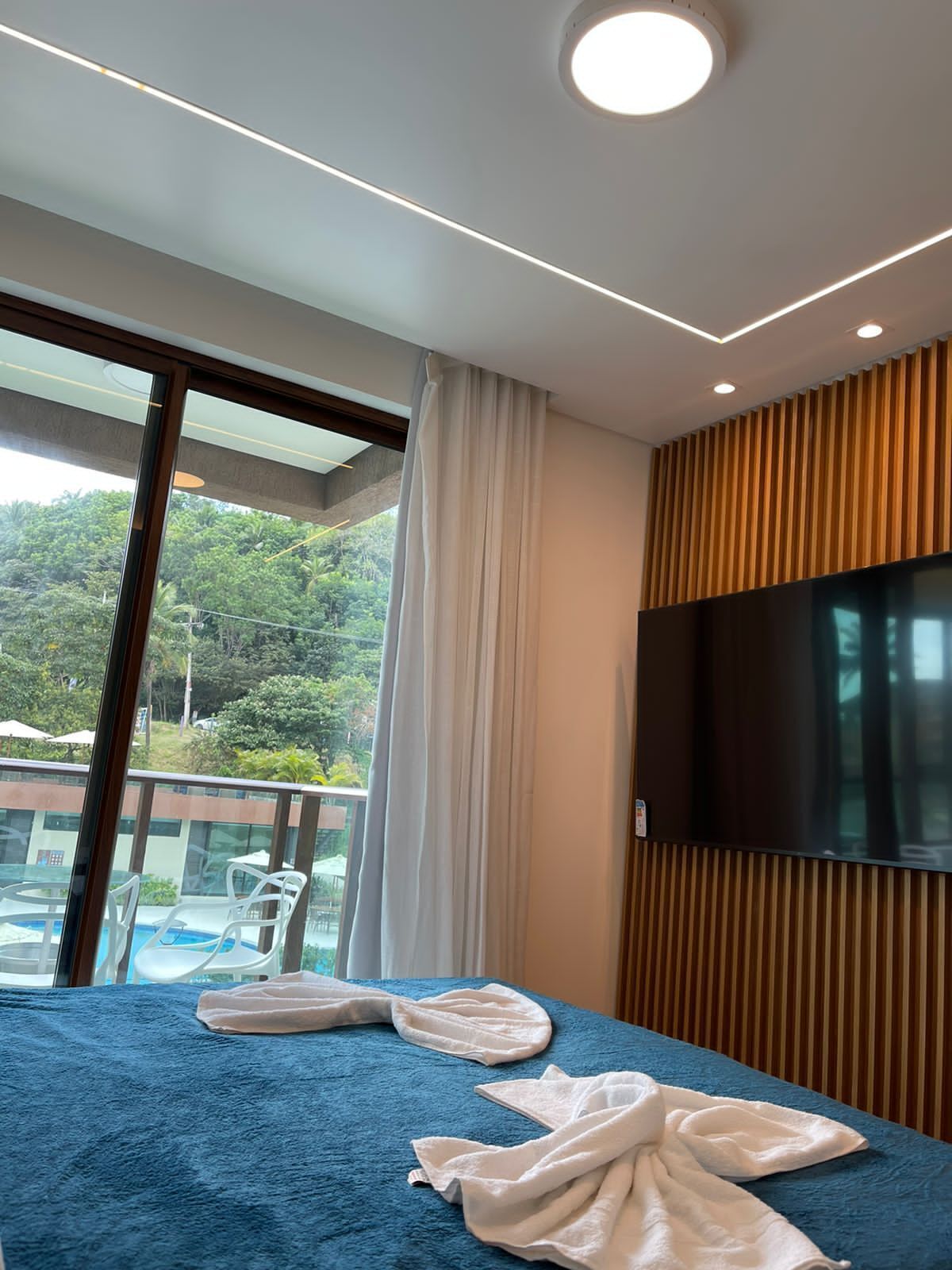 Habitación de hotel con un ventanal grande, una cama con cubierta azul, un televisor montado en la pared y vistas a una piscina y vegetación.