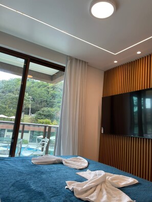A hotel room with a large window, a bed with a blue cover, a TV mounted on the wall, and a view of a pool and greenery outside.