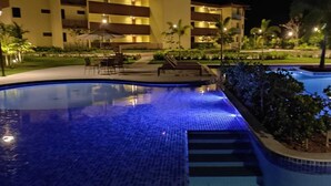A nighttime swimming pool with tiled edges, surrounded by a landscaped area with chairs and a building in the background.