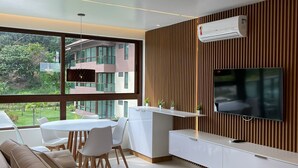 A modern living room with a large TV, a sofa, a dining table, and a view of a building outside.