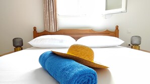 A neatly made bed with a blue towel and a straw hat, a wooden headboard, and a window with curtains. - Oasis Apartments (Santorini)