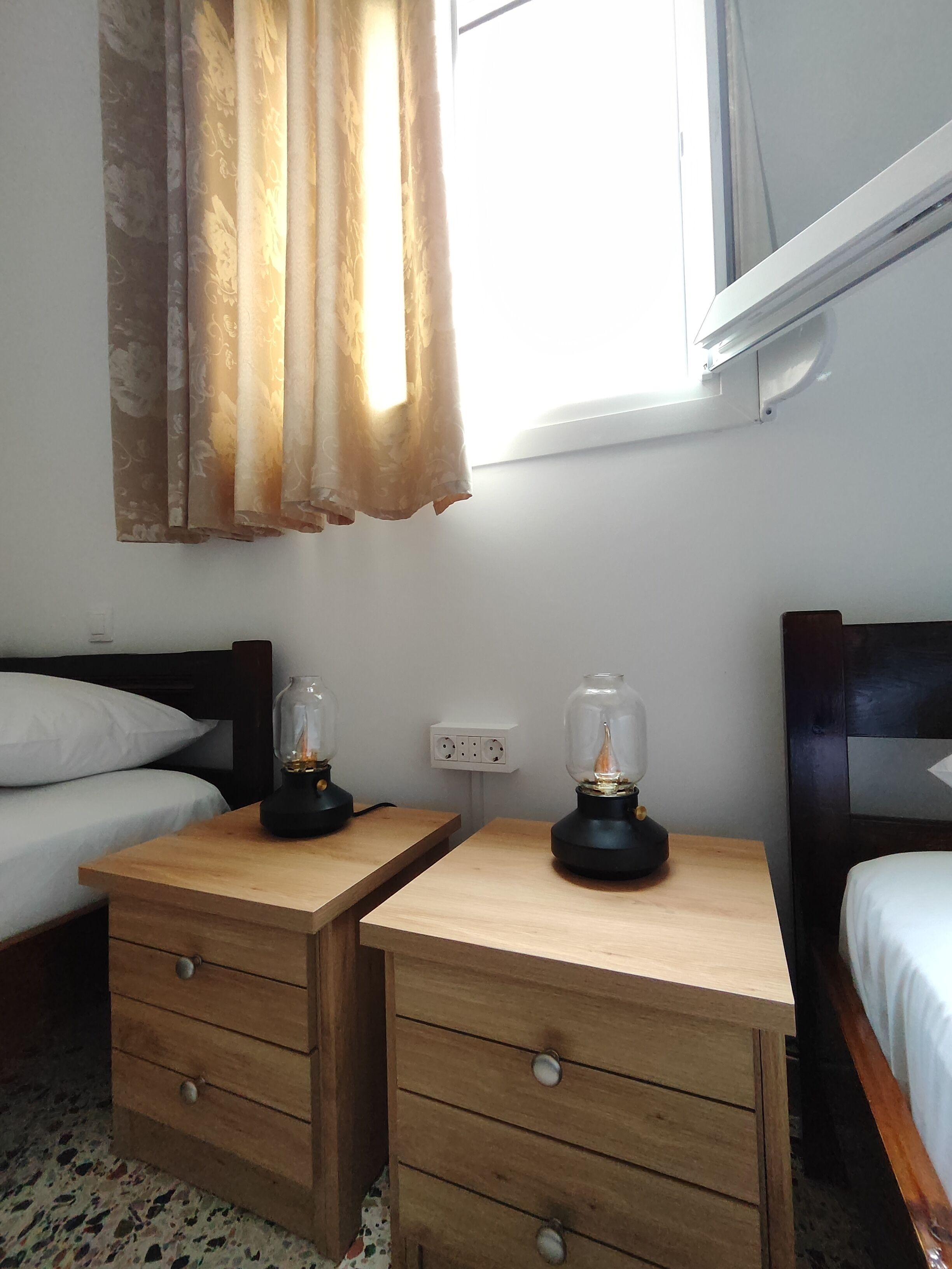 Une chambre à coucher avec deux tables de chevet en bois, chacune équipée d’une lampe, un lit avec une literie blanche et une fenêtre avec des rideaux.