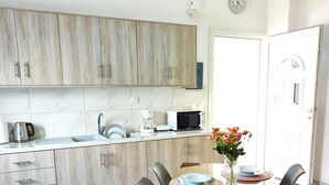 A modern kitchen with wooden cabinets, a microwave, and a sink. There is a dining table with chairs and a vase of flowers. - Oasis Apartments (Santorini)