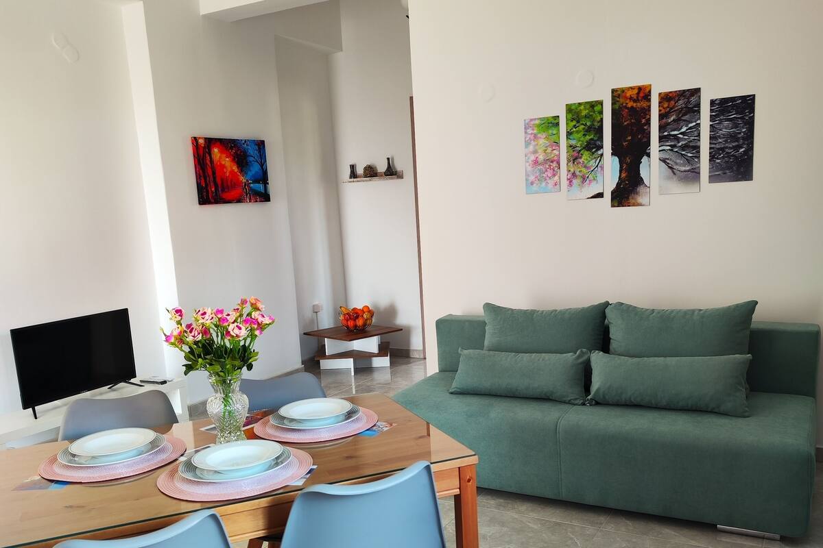 A modern living room with a green sofa, a dining table set with plates and a vase of flowers, and a TV on the left.