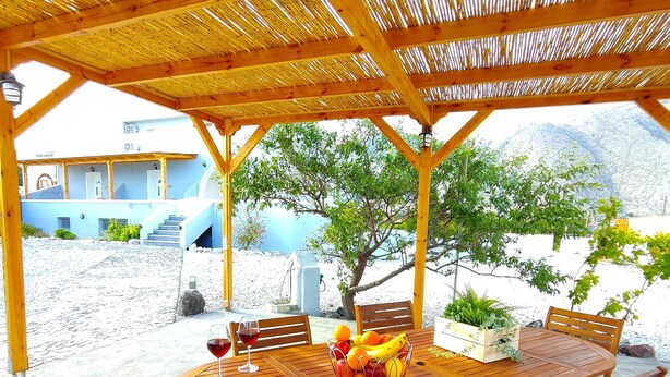 A wooden table with chairs under a thatched roof, surrounded by a sandy area with a view of a building and trees. - Oasis Apartments (Santorini)