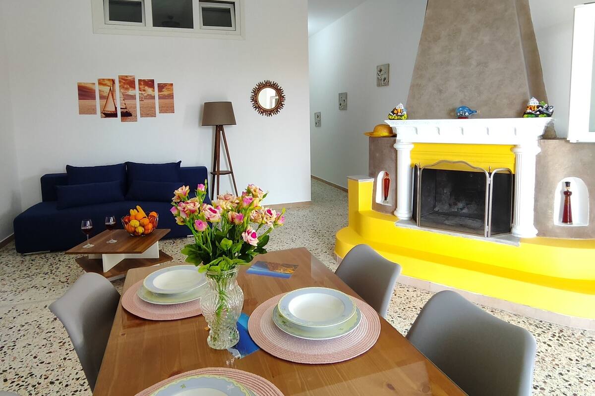 A dining area with a wooden table set for four, a vase with flowers, and a fireplace with colorful decorations.