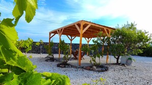 A wooden pergola structure with a gravel ground, surrounded by greenery and a stone wall. - Oasis Apartments (Santorini)