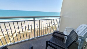 A balcony with a view of the beach, featuring a railing, a wicker chair, and a white plastic chair.