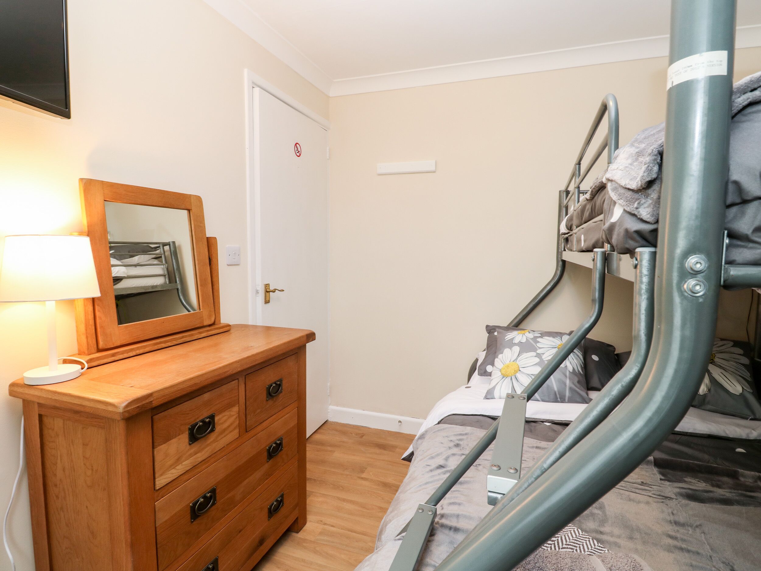 A bunk bed with a metal frame, a wooden dresser with drawers, a bedside lamp, and a door with a no entry sign.