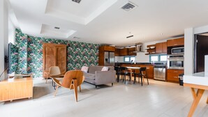 A modern living room with a sofa, wooden chair, and a dining area with a table and chairs. A kitchen with wooden cabinets and appliances is visible in the background. - Casa Azul (Panama City)
