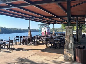 A covered outdoor seating area with tables and chairs, a stone pillar, and a view of a lake.