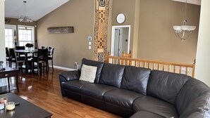 Ein Wohnzimmer mit einem Ecksofa, einem Holzesstisch und Stühlen. Eine Treppe mit Geländer führt zu einer oberen Ebene.