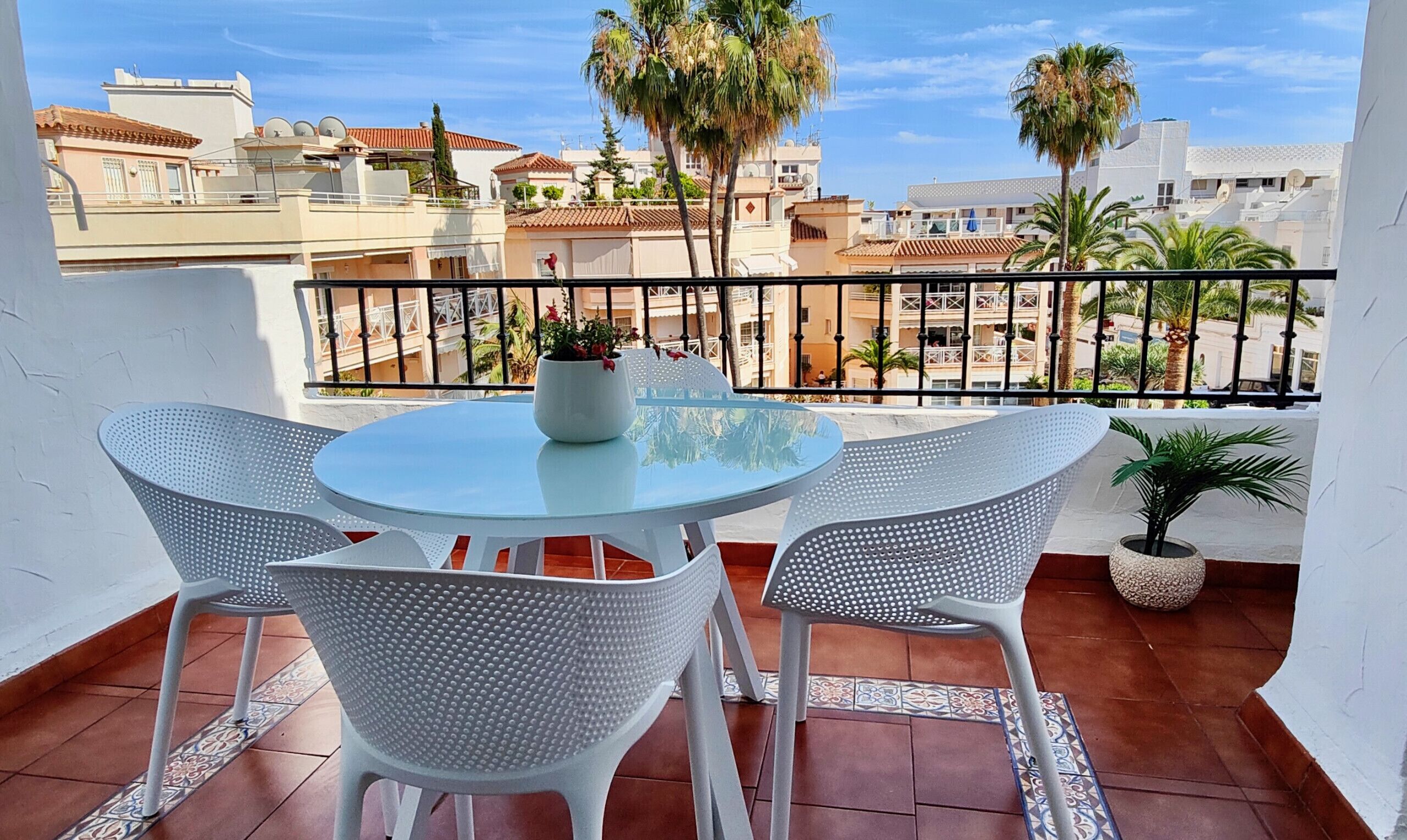 Un balcon avec une table ronde et quatre chaises blanches, un pot de fleurs et une vue sur des immeubles et des palmiers.
