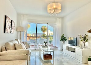 A modern living room with a sofa, a coffee table, a TV, and a balcony with a view of palm trees and buildings. - AP387 ALCAZABA SOUTH VIEW (Nerja)
