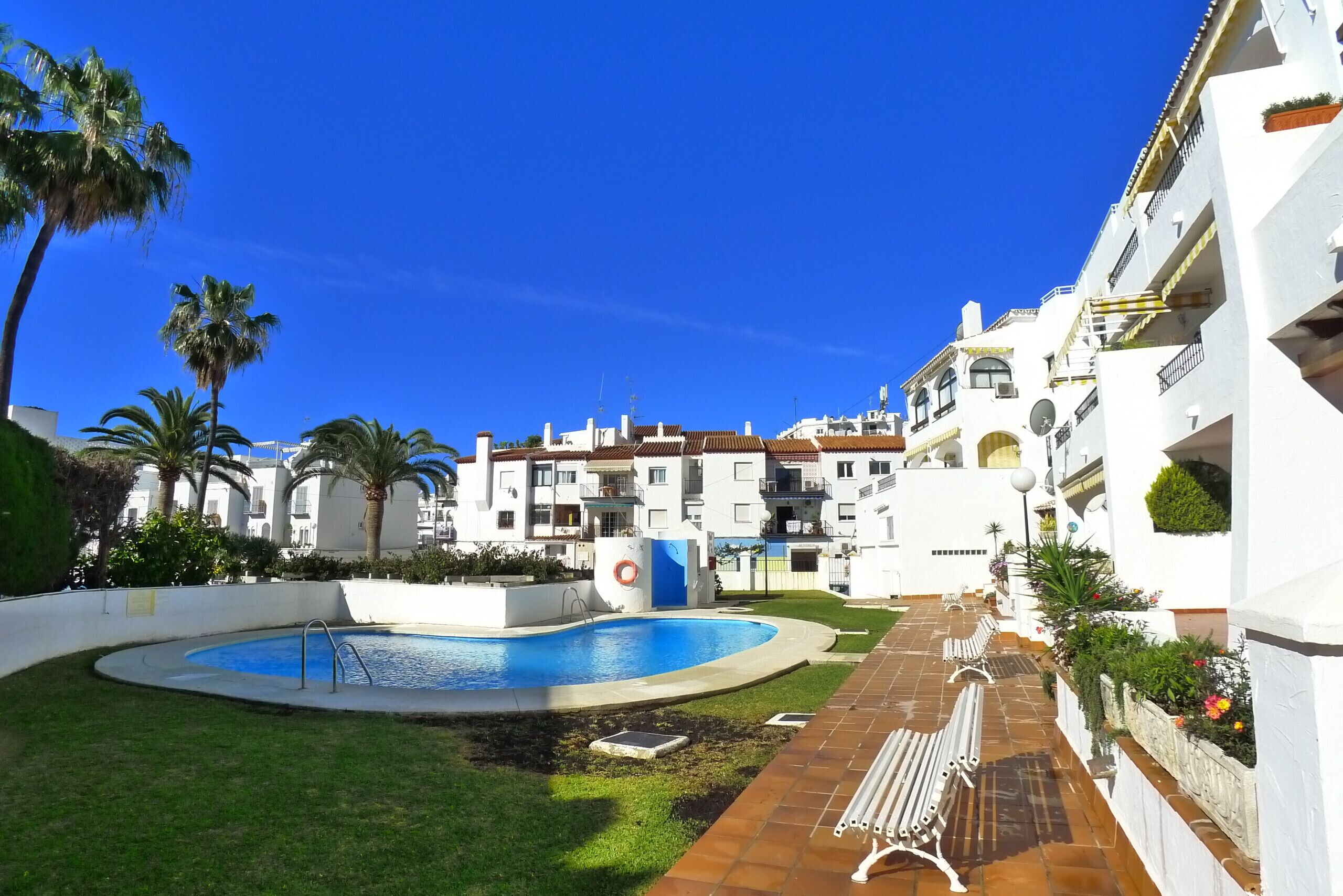 Zona de piscina con una piscina azul, rodeada por un edificio blanco con balcones y un cielo azul despejado.