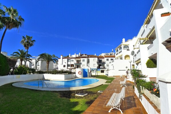 A pool area with a blue pool, surrounded by a white building with balconies and a clear blue sky. - AP387 ALCAZABA SOUTH VIEW (Nerja)