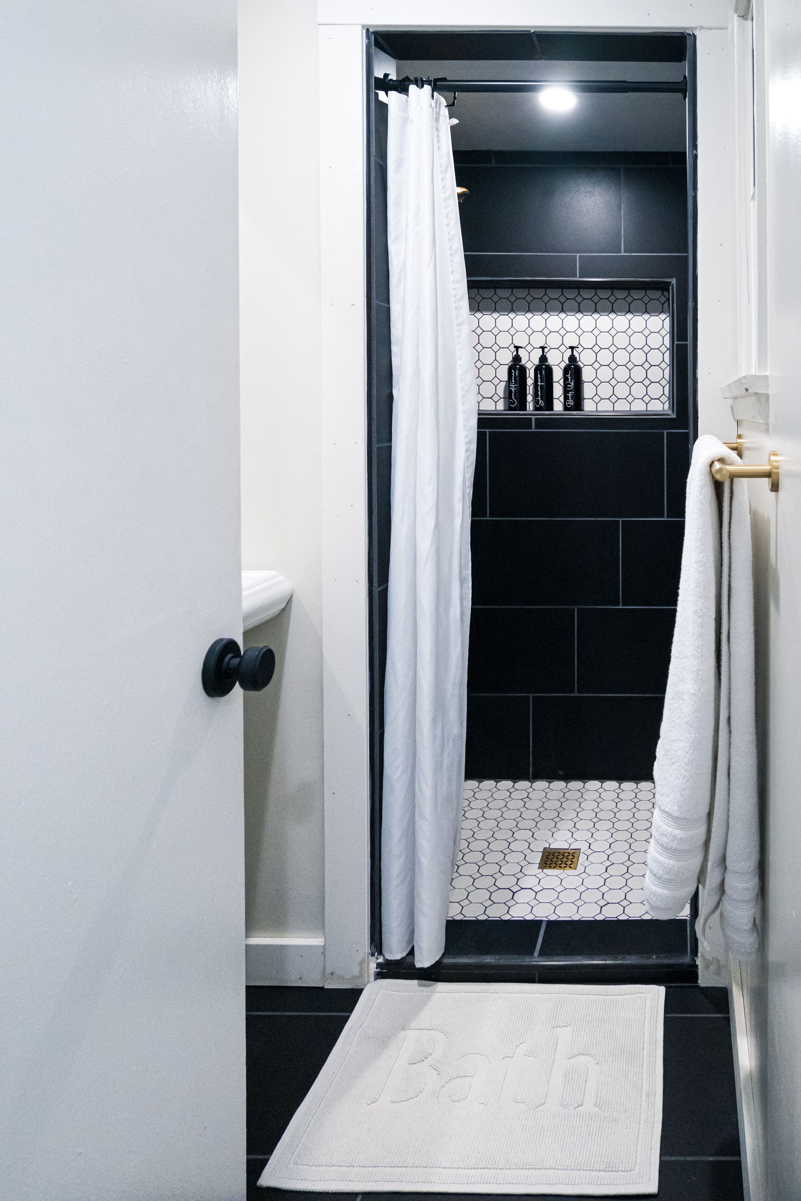 Et badeværelse med en hvid dør, et bruseområde med sorte fliser og en hvid badeværelsesmåtte med ordet "Bath".