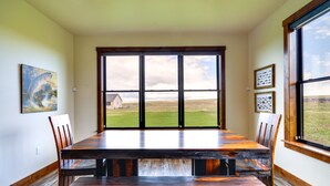 Uma sala de jantar com uma grande mesa de madeira e cadeiras, vista para um campo gramado através de amplas janelas e obras de arte nas paredes.