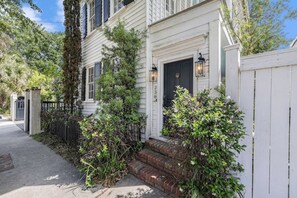 Traditional two-story house with a brick entrance and a small porch area, surrounded by lush greenery and trees.