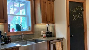 A kitchen with wooden cabinets, a window, a chalkboard with 'Summer 2023' written on it, and a door with a towel hanging on it. - CREEKSIDE RETREAT 5 miles from Cooperstown, NY. (Hartwick)
