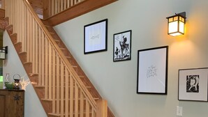 A wooden staircase with a handrail, a wall with framed artwork, and a wall-mounted light fixture. - CREEKSIDE RETREAT 5 miles from Cooperstown, NY. (Hartwick)