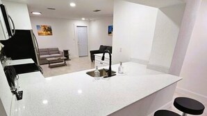 Eine moderne Küche mit weißer Arbeitsplatte, schwarzen Barhocker und einem Essbereich im Hintergrund.
