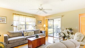 A spacious living room with a grey sofa, a wooden coffee table, and a beige sofa. There is a ceiling fan, a large window with curtains, and a door leading to a balcony. - Complimentary Golf Cart! Home in The Villages (The Villages)