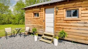 A wooden cabin with a front door, two windows, a small porch with steps, and a table with chairs.