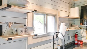 A kitchen with wooden walls, a window, a coffee maker, mugs, a paper towel dispenser, and a fire extinguisher.