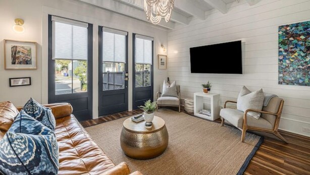 A spacious living room with a leather sofa, a round coffee table, and a chandelier. There are three large windows and a flat-screen TV mounted on the wall. - Downtown Charleston! One block to King St. Free parking for two vehicles (Charleston)