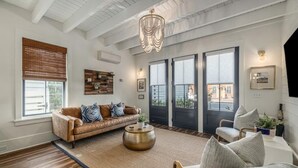 A spacious living room with a brown leather sofa, a round coffee table, and a grey armchair. There are two large windows with blinds, a chandelier, and a wall-mounted TV. - Downtown Charleston! One block to King St. Free parking for two vehicles (Charleston)