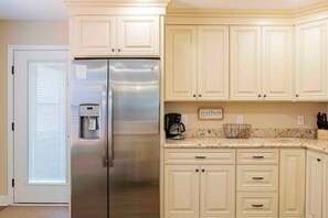 A kitchen with light-colored cabinets, a stainless steel refrigerator, a coffee maker, and a 'gather' sign on the wall.