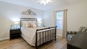 Een slaapkamer met een bed, nachtkastjes, een plafondventilator, een raam met gordijnen en een gestreept stoeltje.