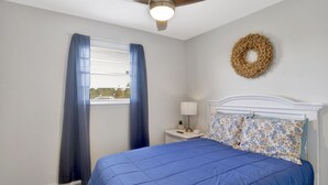 A bedroom with a bed, a ceiling fan, a window with curtains, a nightstand with a lamp, and a wall wreath. - Cozy Coleman-Families- Near Beaches, Airport & PCC (Pensacola)